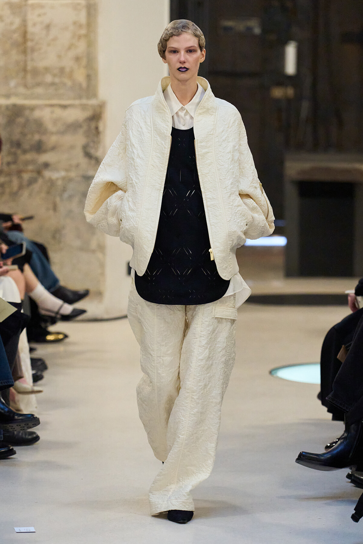Model on a runway wearing a cream quilted suit with oversized jacket over a black top, white shirt, and matching pants; audience seated along the catwalk behind.