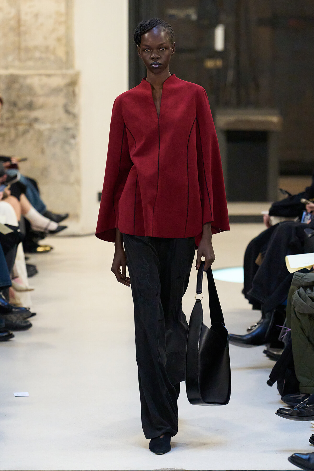 Model walks a runway in a deep red cape-style top, black wide-leg pants, and a large black tote.