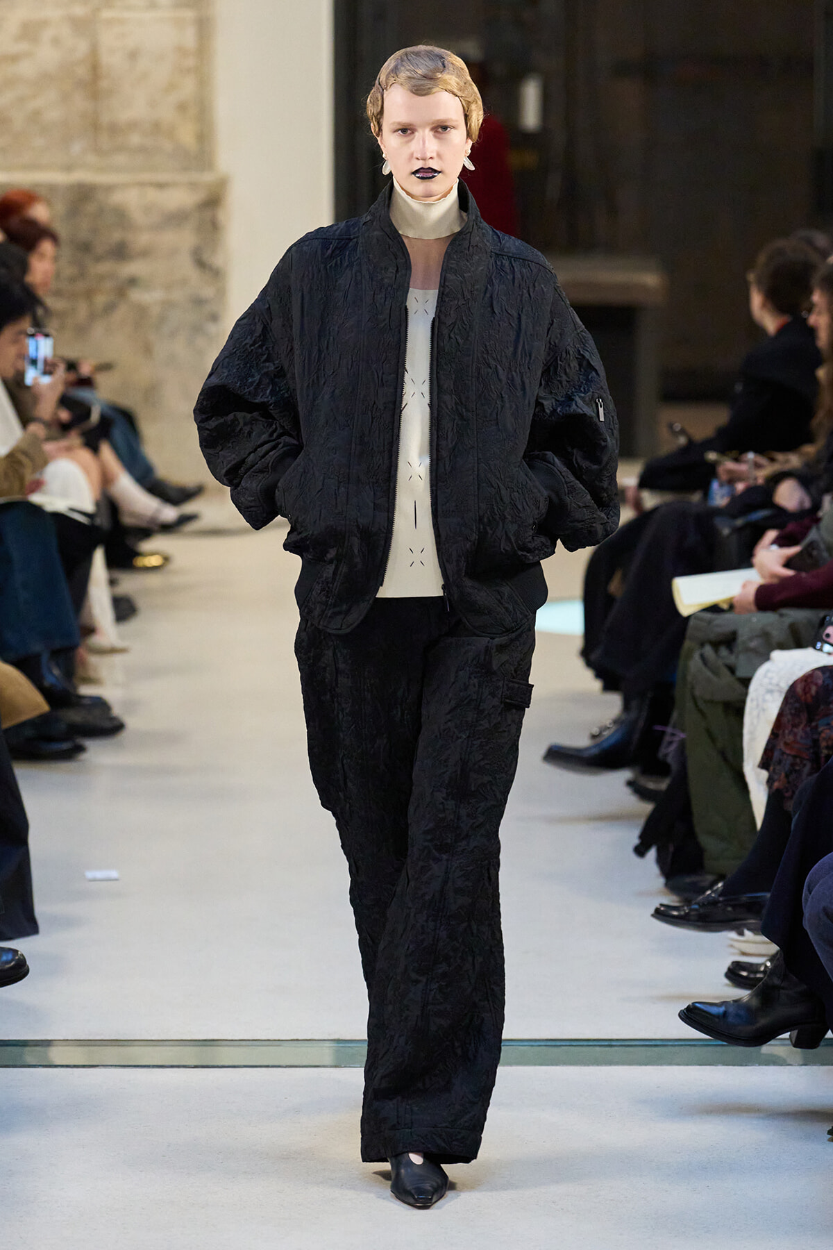 Model walks a runway in an oversized black textured quilted jacket and matching pants with short blond hair and dark lipstick, audience on sides.