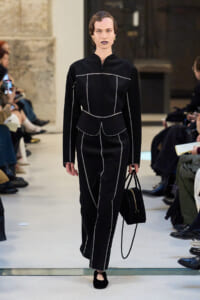 Model walks a runway in a black tailored outfit with white stitching, carrying a black handbag as spectators watch on both sides.
