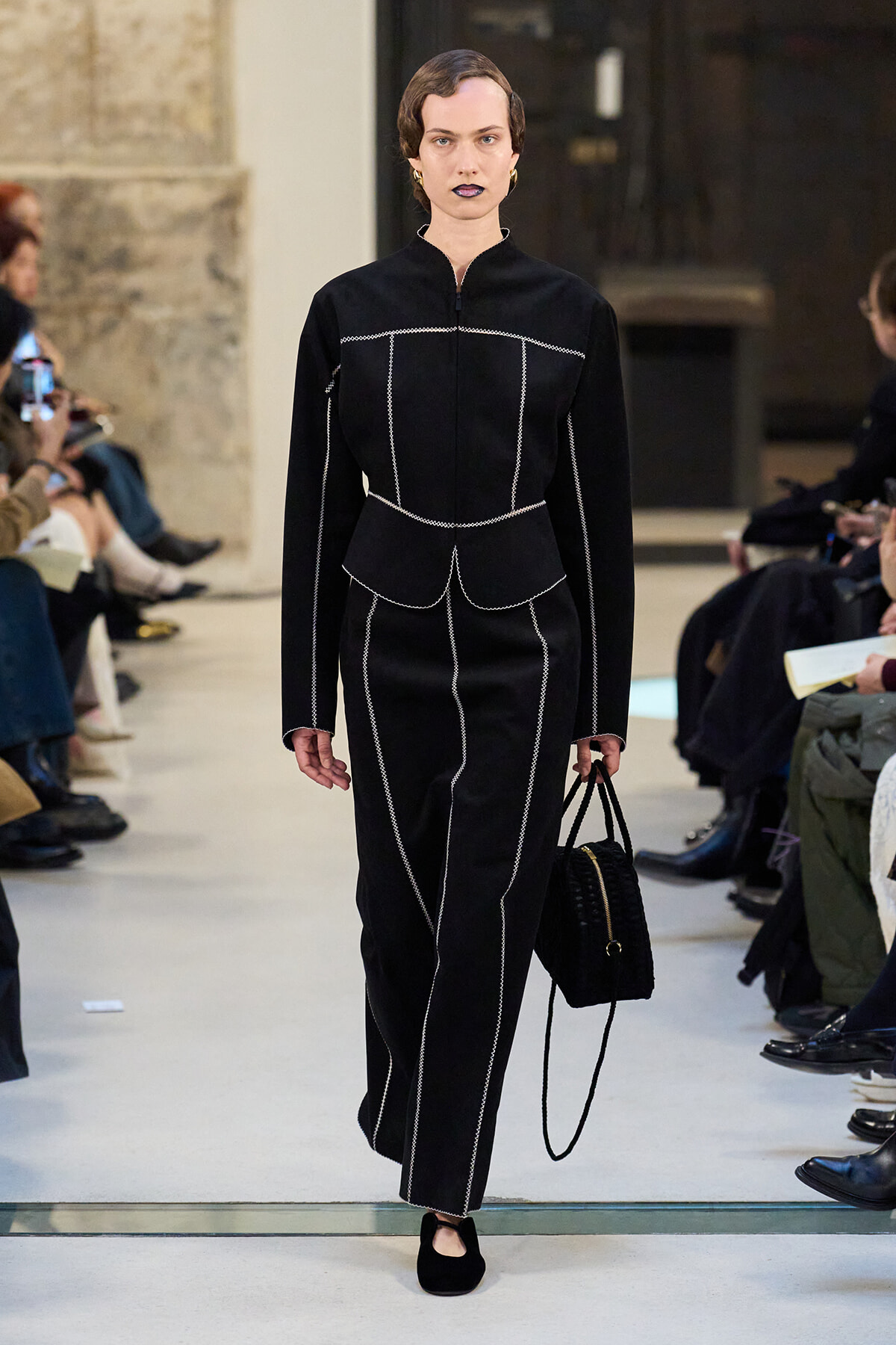 Model walks a runway in a black tailored outfit with white stitching, carrying a black handbag as spectators watch on both sides.