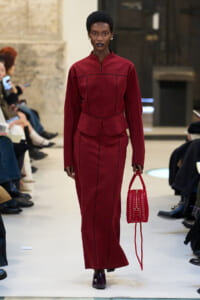 Model walking a runway in a burgundy, structured long-sleeve dress with a peplum and matching long skirt, carrying a red woven handbag parallel to her side.