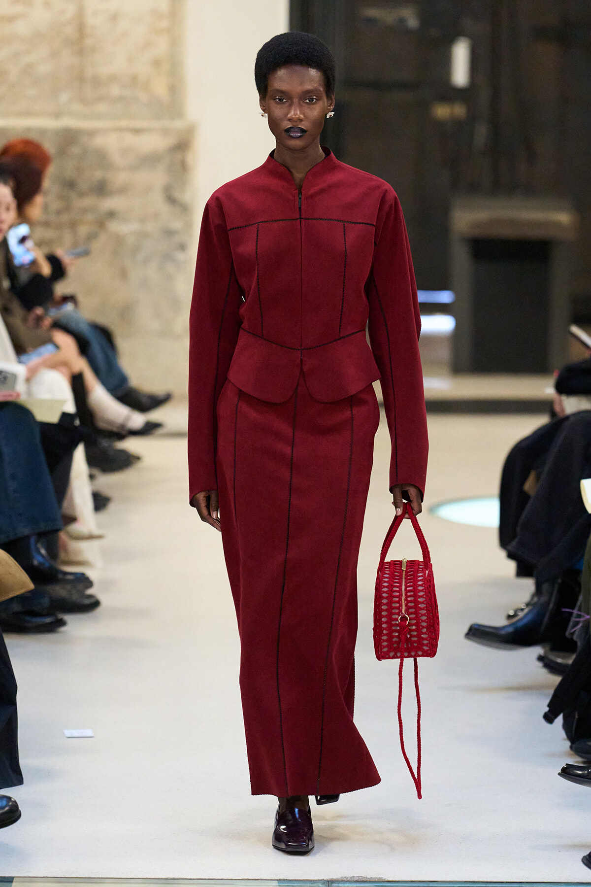 Model walking a runway in a burgundy, structured long-sleeve dress with a peplum and matching long skirt, carrying a red woven handbag parallel to her side.