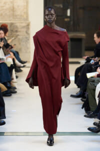 Runway model in a burgundy off‑the‑shoulder dress with flared sleeves, black lipstick, and gold earrings, walking the catwalk surrounded by seated audience.