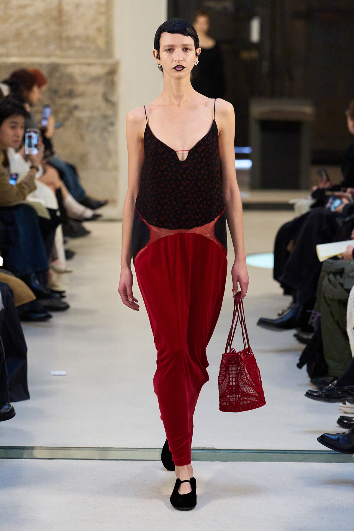 Model walks the runway in a black and red velvet outfit with a red crochet handbag.