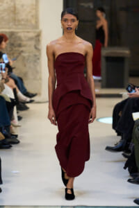 Model walks a burgundy strapless gown with layered draped ruffles down the skirt on a fashion runway.