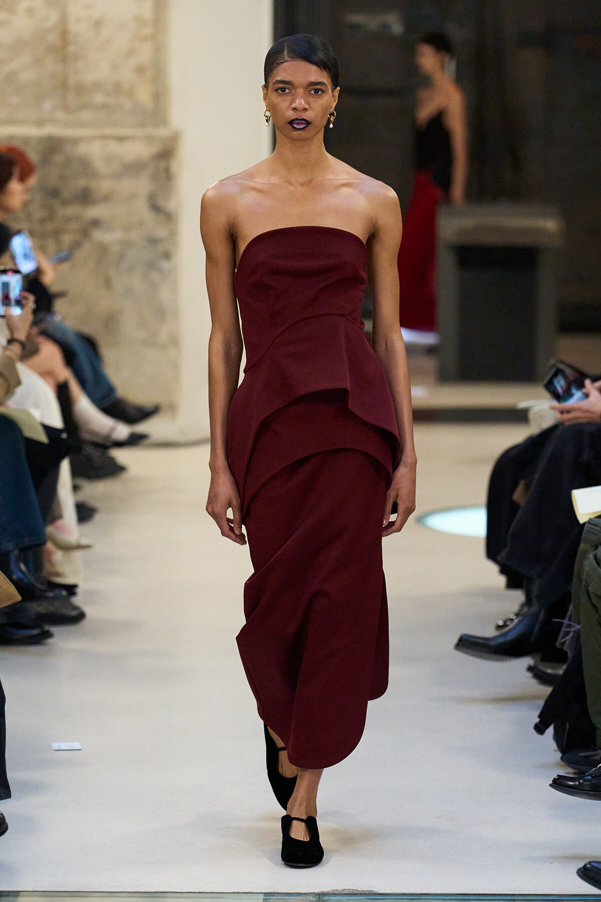 Model walks a burgundy strapless gown with layered draped ruffles down the skirt on a fashion runway.