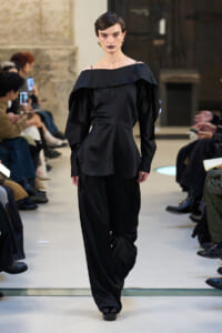 Model walking a black off-the-shoulder outfit with wide legs on a runway, audience seated on both sides watching.