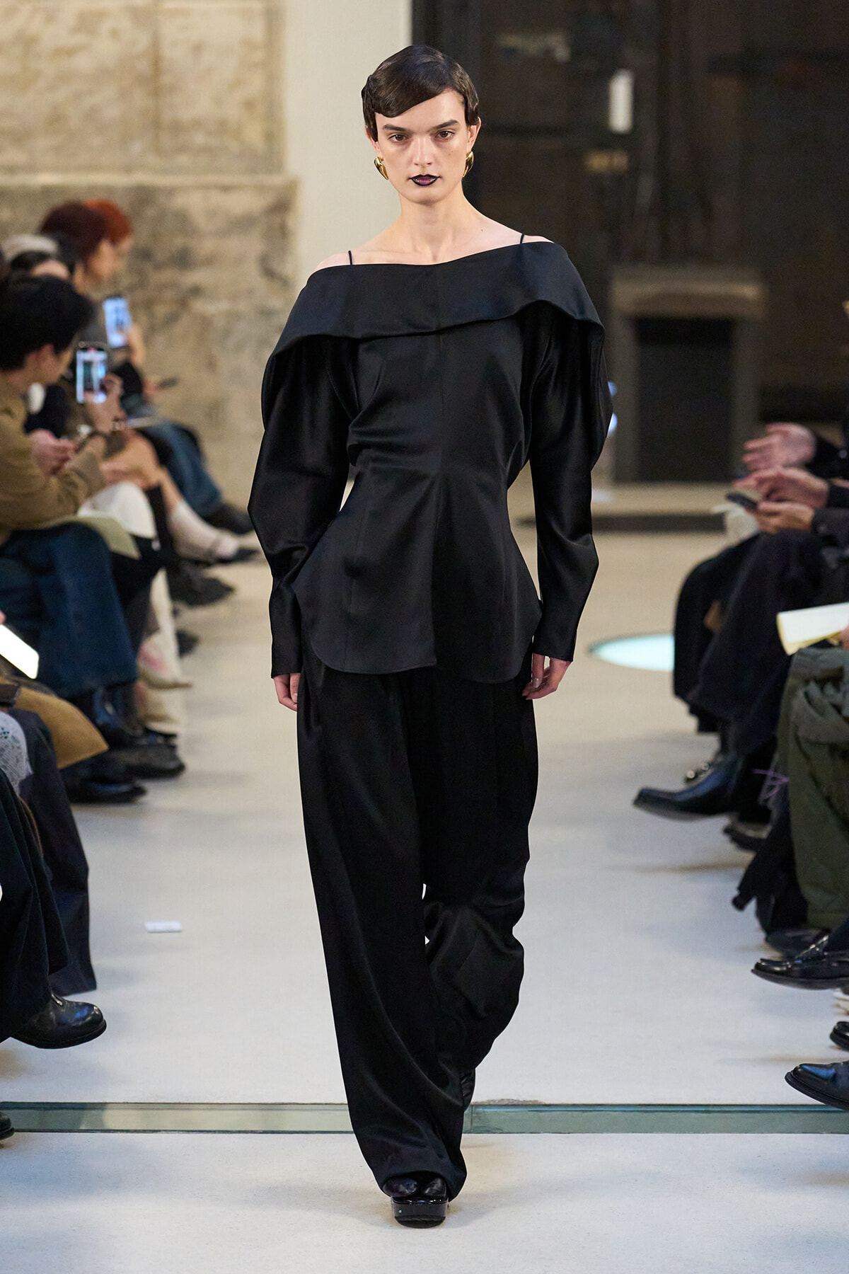 Model walking a black off-the-shoulder outfit with wide legs on a runway, audience seated on both sides watching.