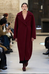 Model walks a runway in a long burgundy coat with wide lapels, paired with dark boots and bold lipstick.