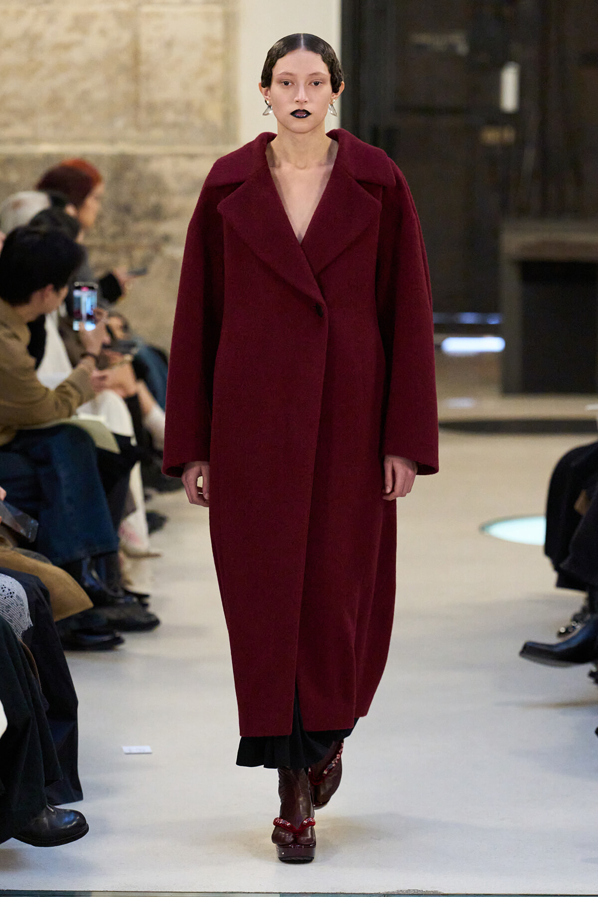 Model walks a runway in a long burgundy coat with wide lapels, paired with dark boots and bold lipstick.