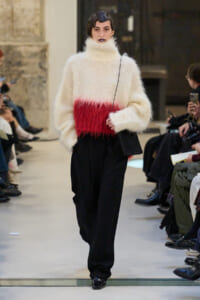 Model walking a runway in a cream fuzzy turtleneck sweater with a red band, black wide-leg pants, and a small black crossbody bag.