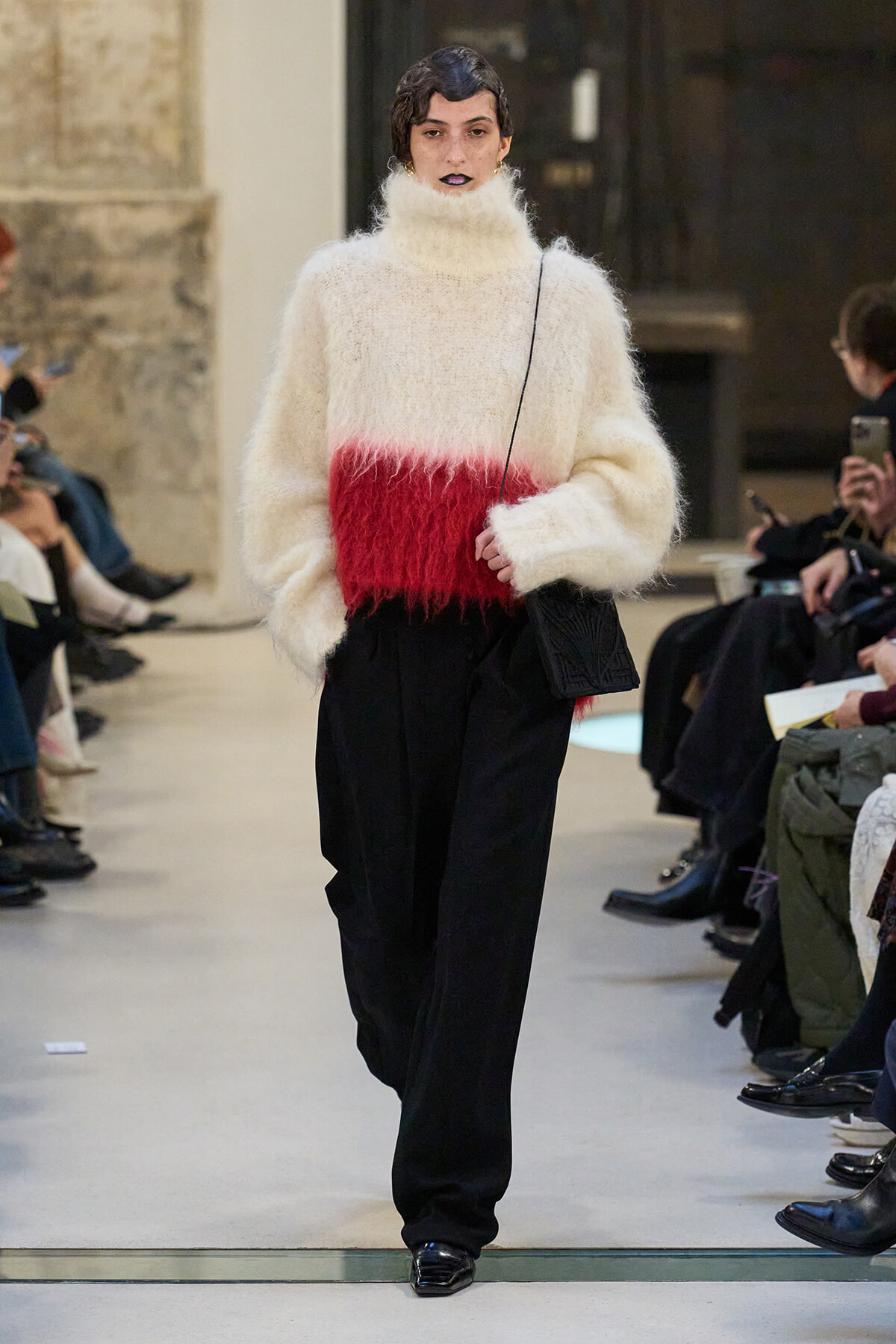 Model walking a runway in a cream fuzzy turtleneck sweater with a red band, black wide-leg pants, and a small black crossbody bag.