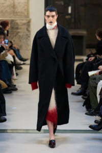 Model walking the runway in a long black coat with red fuzzy cuffs and hem, paired with a white sweater and red scarf.