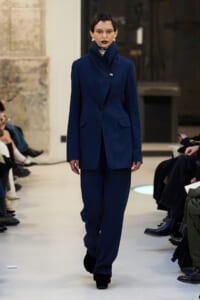 Model walks a runway wearing a tailored navy blue pantsuit with a high-collared blazer and matching wide-leg trousers, with black lipstick and pearl earrings.
