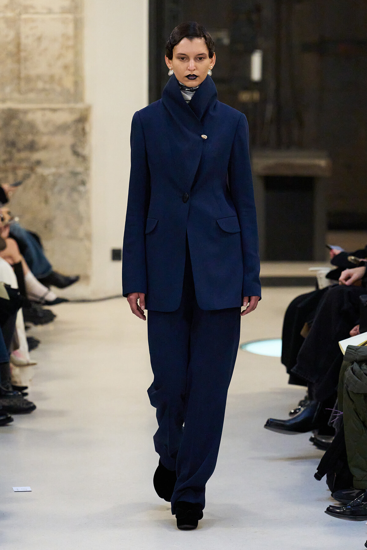 Model walks a runway wearing a tailored navy blue pantsuit with a high-collared blazer and matching wide-leg trousers, with black lipstick and pearl earrings.