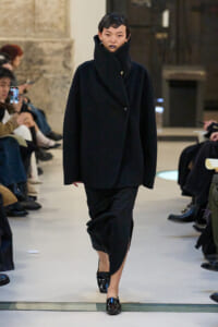 Runway model in an oversized black coat with a tall high collar, black lipstick, walking toward the camera.
