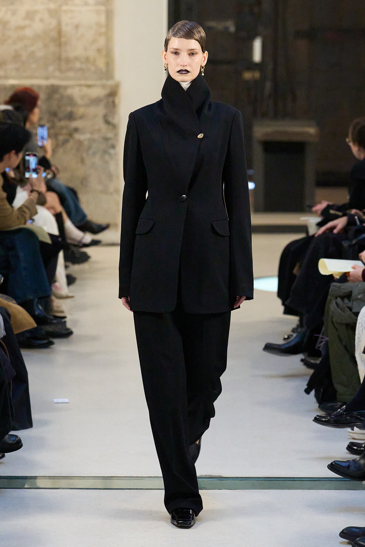 Model walks the runway in a black tailored blazer with a high stand collar and matching wide-leg pants, audience on both sides.