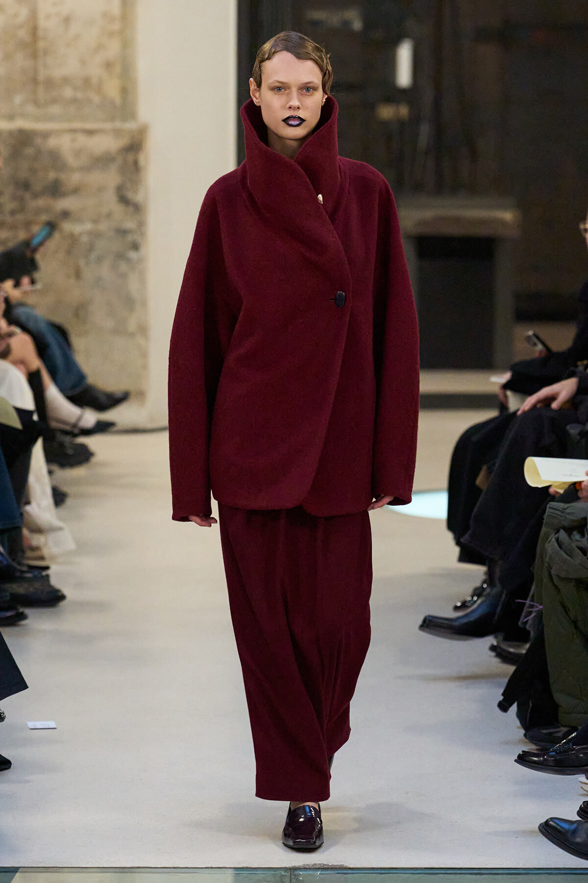 Model walking a runway in a burgundy oversized coat with a high, sculpted collar and matching wide-leg pants.