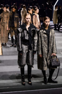 Two models stand on a fashion show runway in metallic patterned coats and black scarves; one holds a quilted handbag.