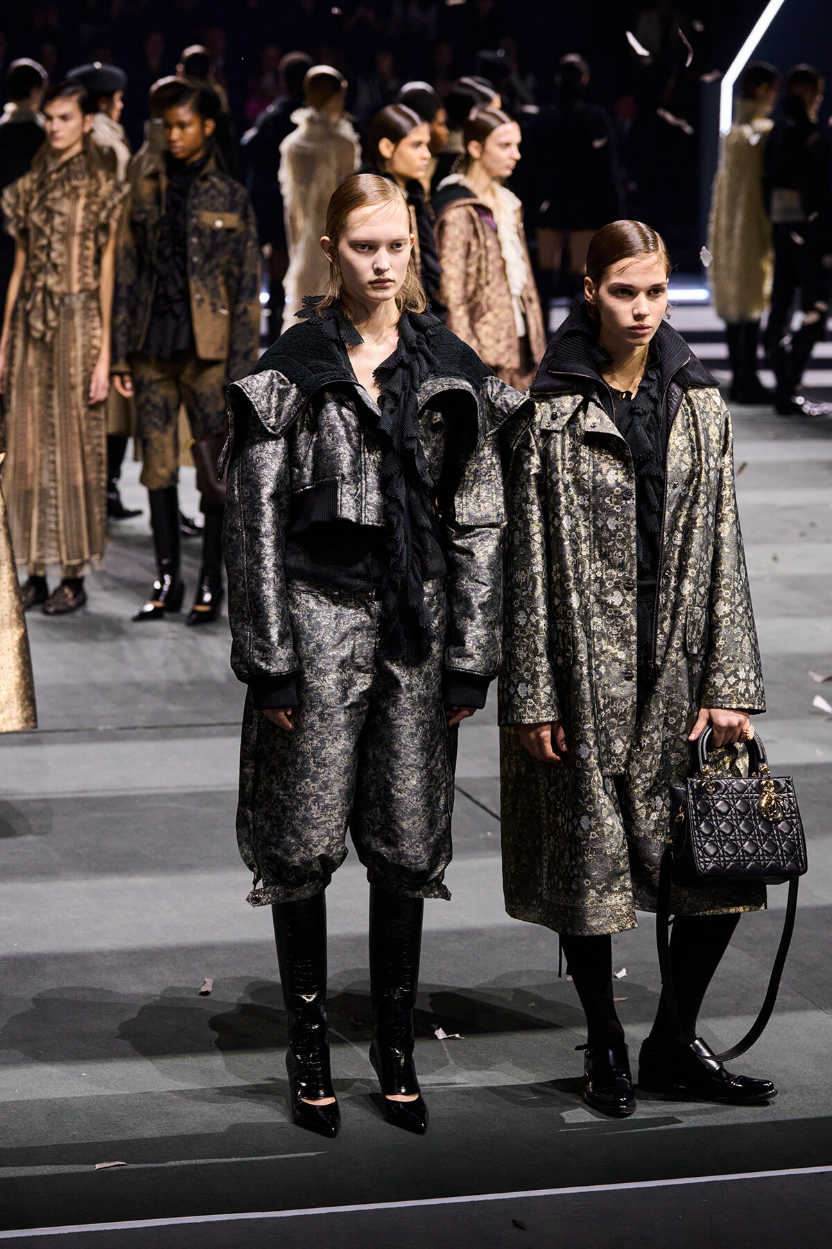 Two models stand on a fashion show runway in metallic patterned coats and black scarves; one holds a quilted handbag.