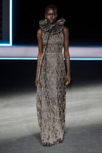 Female model on a runway wearing a floor-length metallic beaded gown with a sculptural ruffled collar.