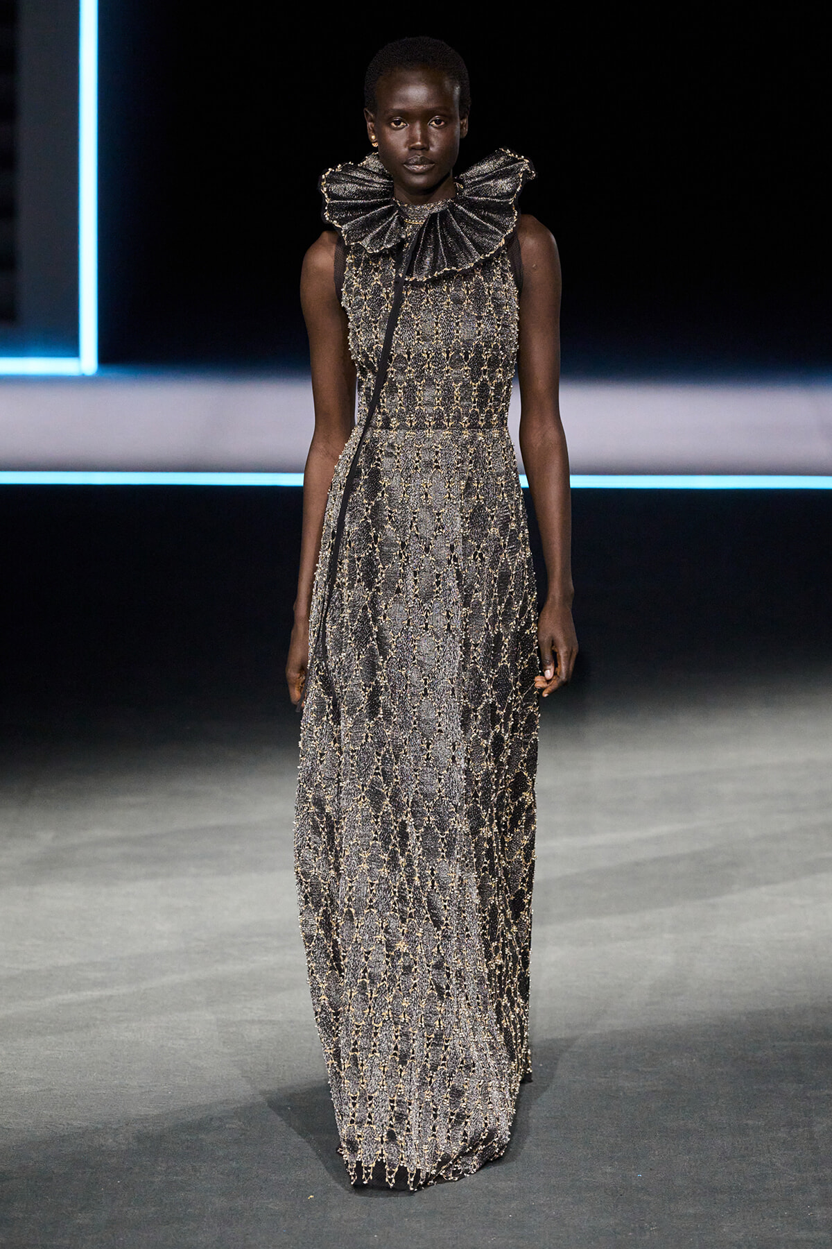 Female model on a runway wearing a floor-length metallic beaded gown with a sculptural ruffled collar.