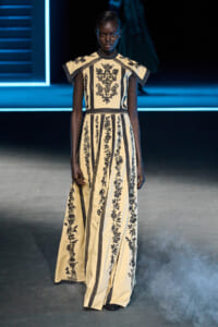 Model on a runway wearing a cream, floor-length gown with black floral embroidery and cape-like shoulders.