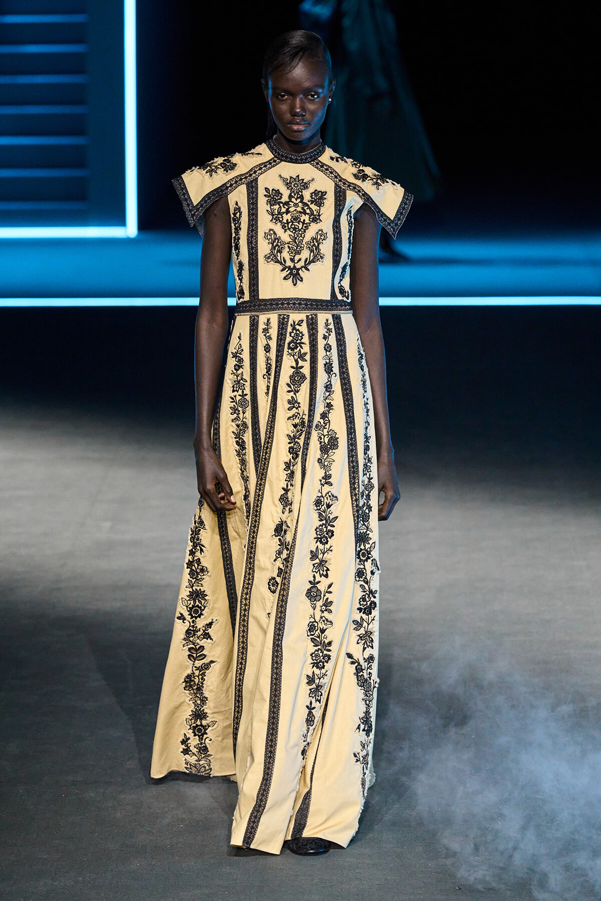 Model on a runway wearing a cream, floor-length gown with black floral embroidery and cape-like shoulders.