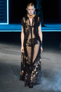 Model on a fashion runway wearing a sheer black embroidered gown with a chunky gold choker on a dimly lit catwalk.