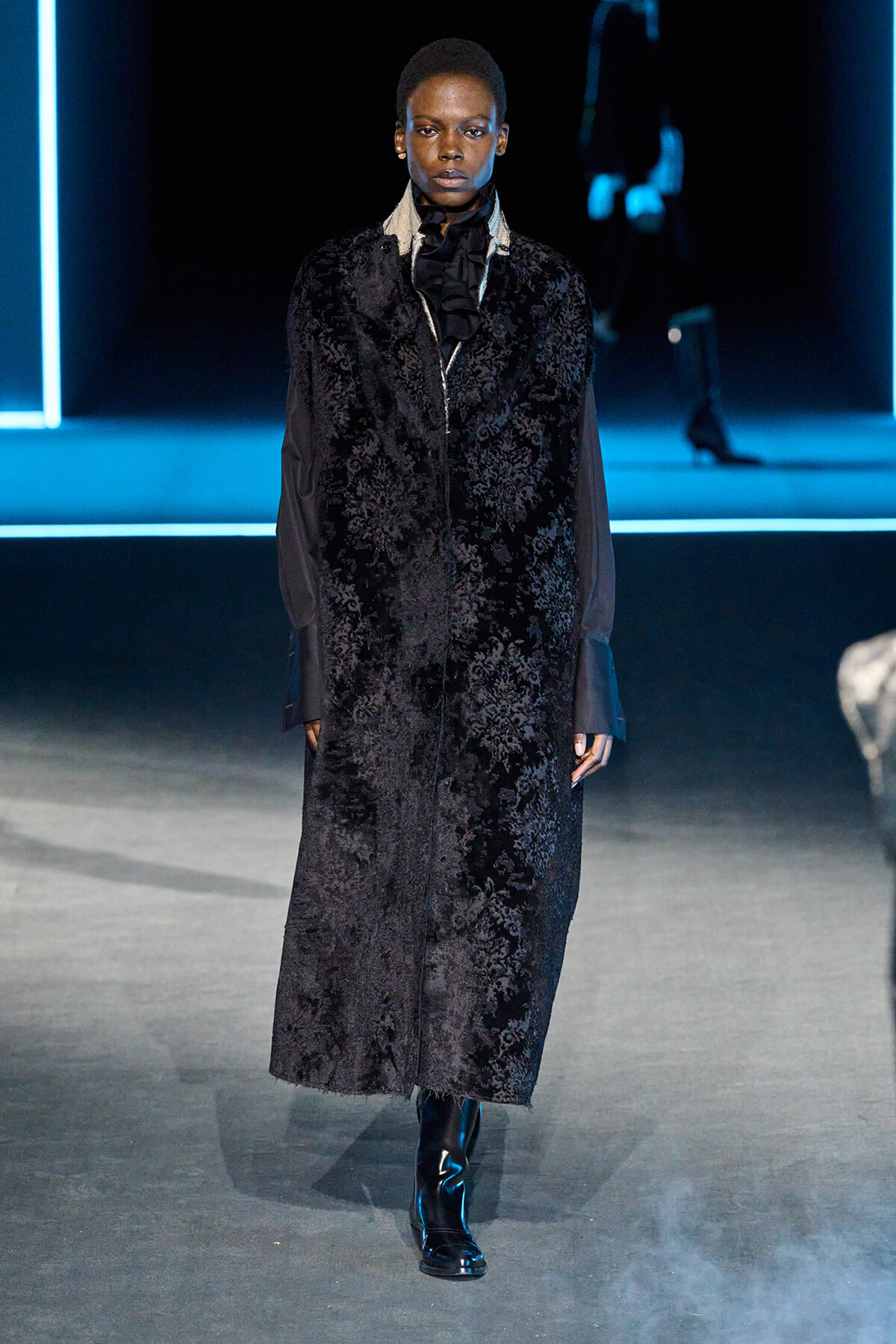 Runway model wearing a floor-length black velvet coat with a subtle floral pattern and a ruffled high-neck blouse, paired with black boots on a neon-lit stage.