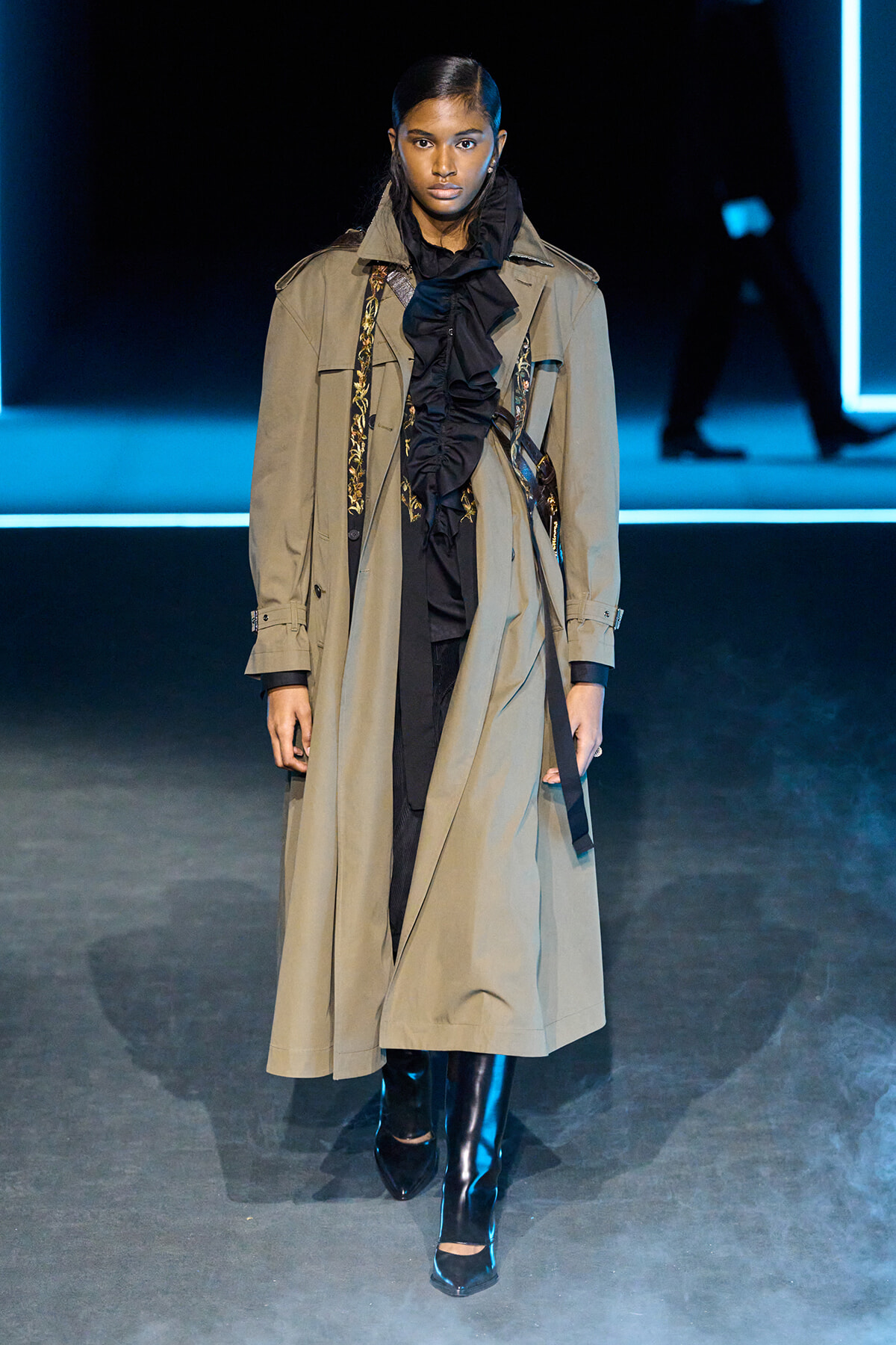 Female model walks a fashion runway wearing a long beige trench coat with a black ruffled scarf and knee-high black boots, blue stage lighting in the background.