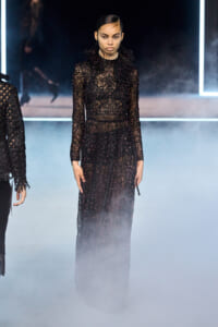 Model in a floor-length black lace sequin gown with a high ruffled collar walking the runway amid fog and neon lights.