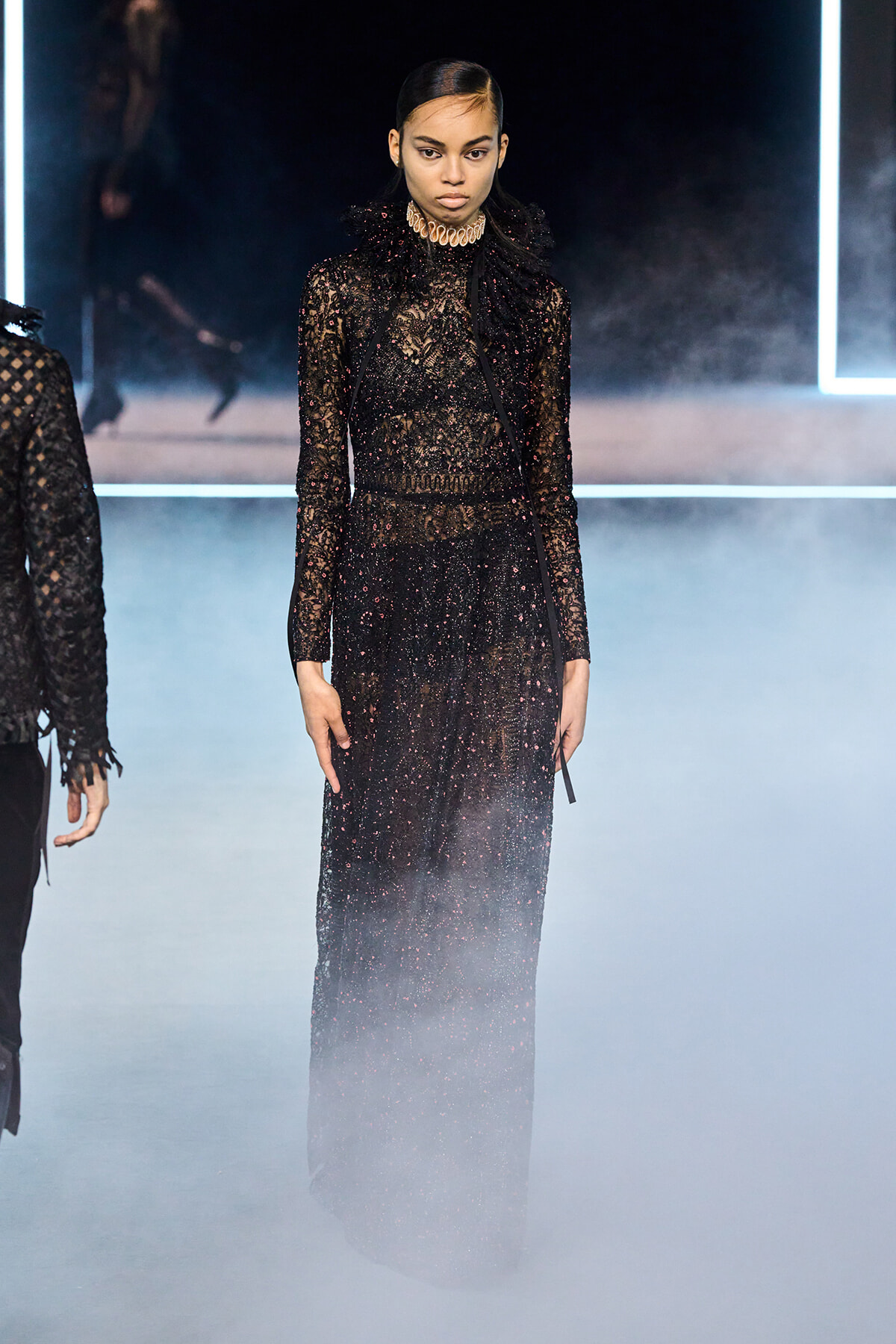 Model in a floor-length black lace sequin gown with a high ruffled collar walking the runway amid fog and neon lights.