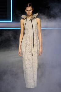 Model on a foggy runway wearing a beige lace sleeveless gown with a dramatic ruffled high collar and a long black cord down the front.