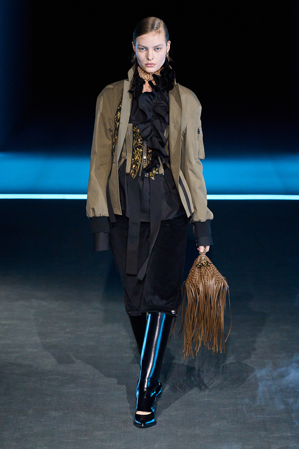 Model walking runway in olive bomber jacket, black ruffled blouse, and black pants, holding a fringed brown suede bag.