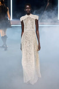 Model wearing ivory lace high-neck, short-sleeve, floor-length gown on a smoky fashion-show runway.