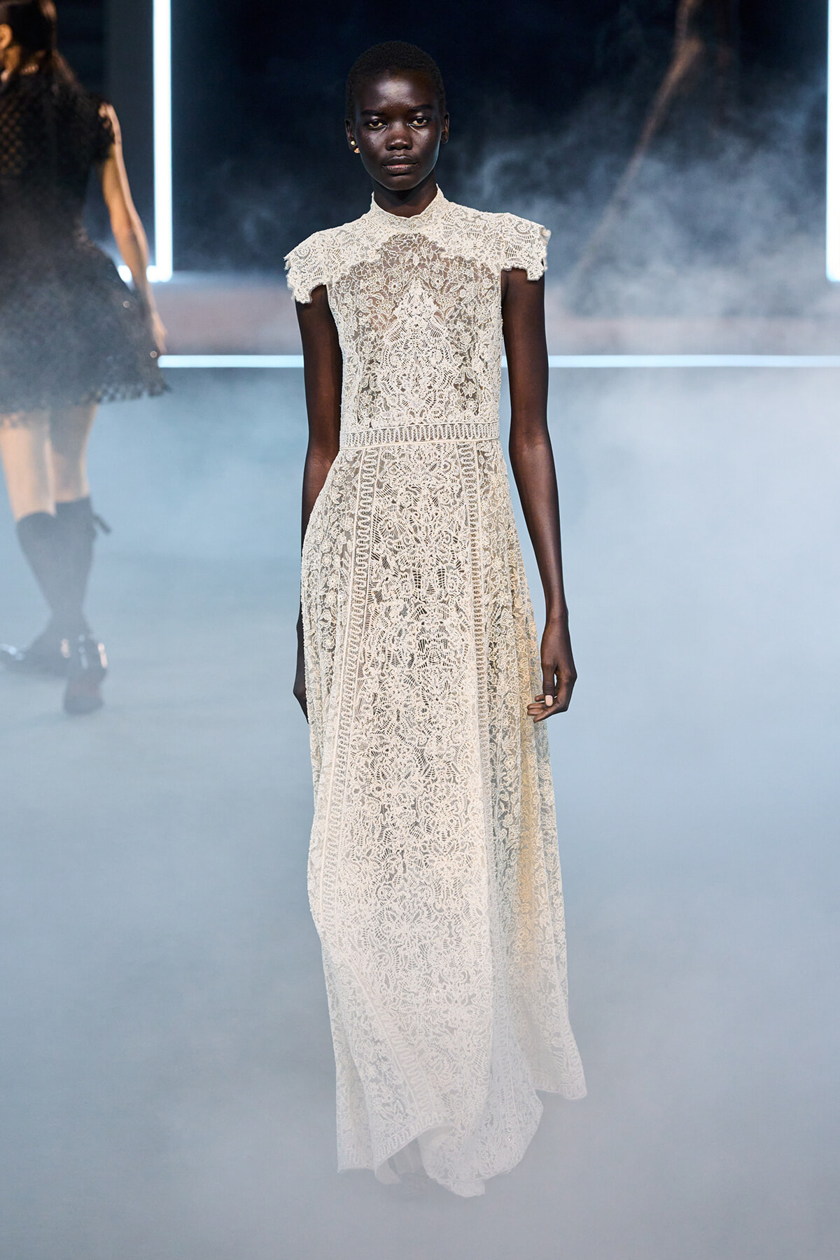 Model wearing ivory lace high-neck, short-sleeve, floor-length gown on a smoky fashion-show runway.