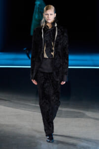 Model walks down the runway in a black velvet suit with a ruffled black blouse and beige accents at the neck.