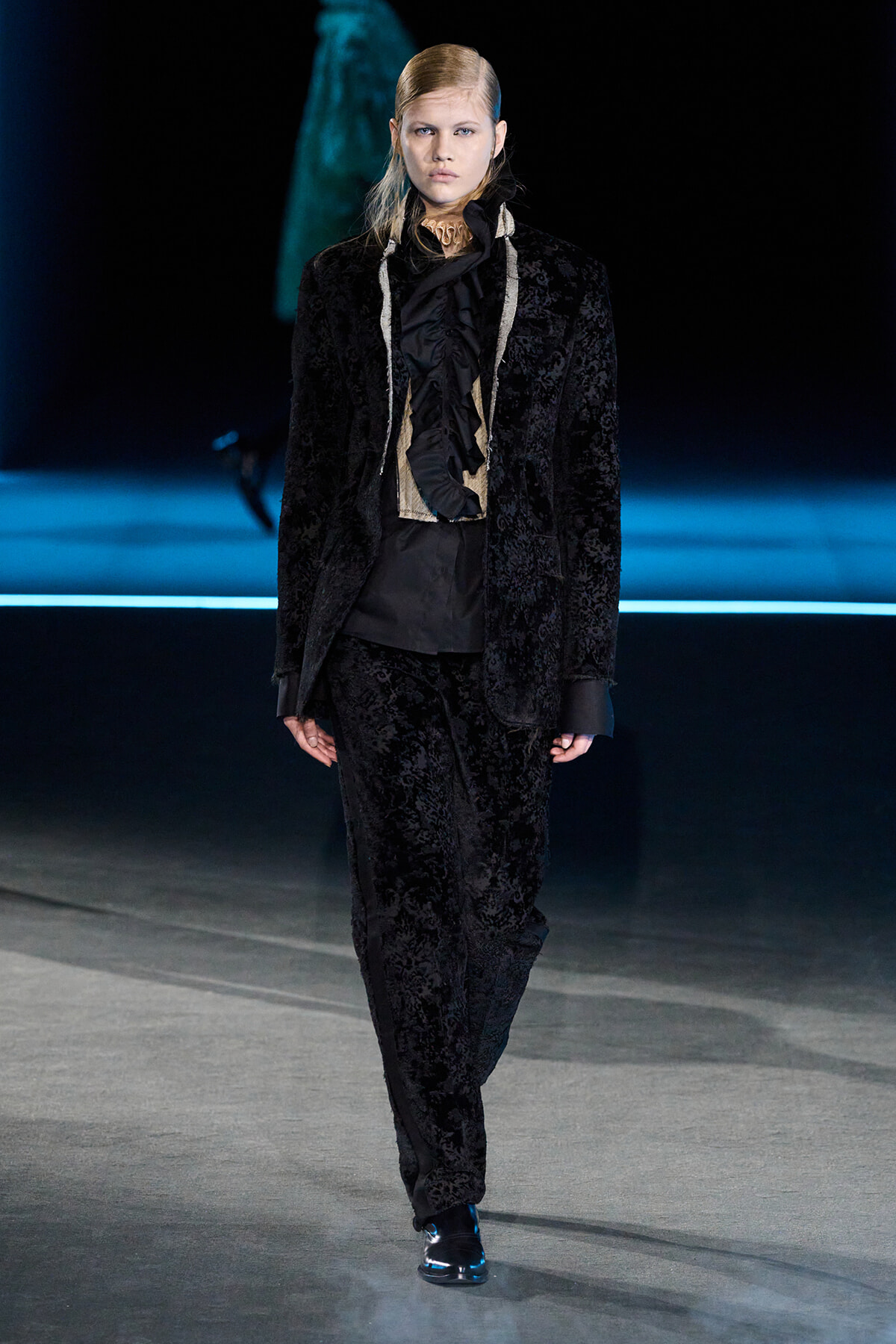 Model walks down the runway in a black velvet suit with a ruffled black blouse and beige accents at the neck.