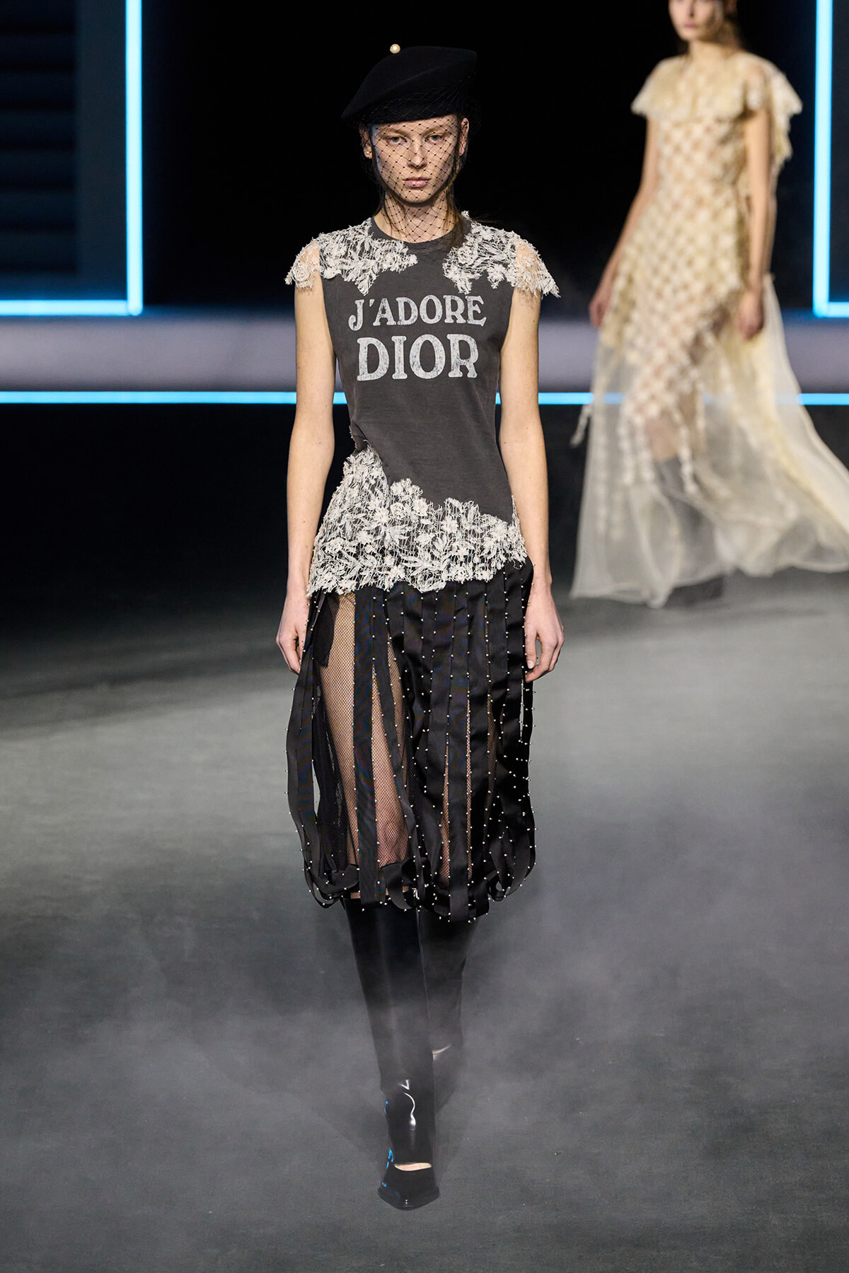 Model on a runway wearing a dark 'J'adore Dior' tee with lace accents and a sheer, beaded skirt with panels, black hat and veil.
