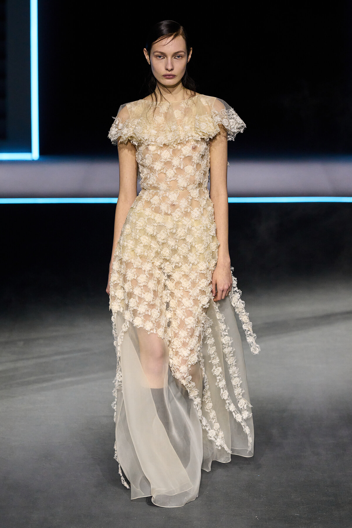 Model walking the runway in a champagne-colored lace gown with sheer overlay and short off-the-shoulder sleeves, at a fashion show.