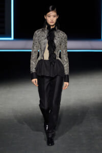 Model walks a fashion runway in a silver-embellished jacket over a black ruffled blouse and black satin pants, black shoes, neon-lit backdrop.