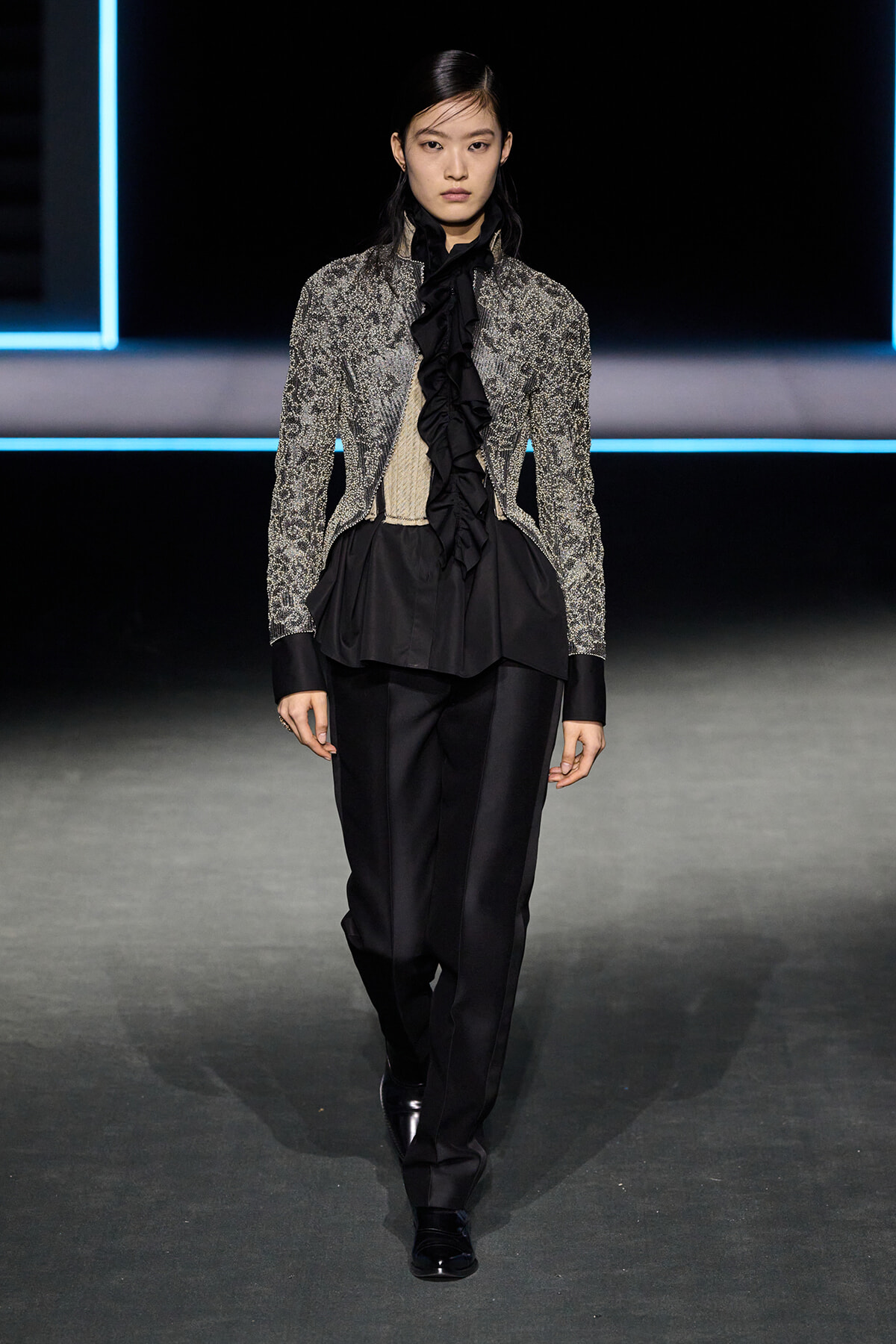 Model walks a fashion runway in a silver-embellished jacket over a black ruffled blouse and black satin pants, black shoes, neon-lit backdrop.