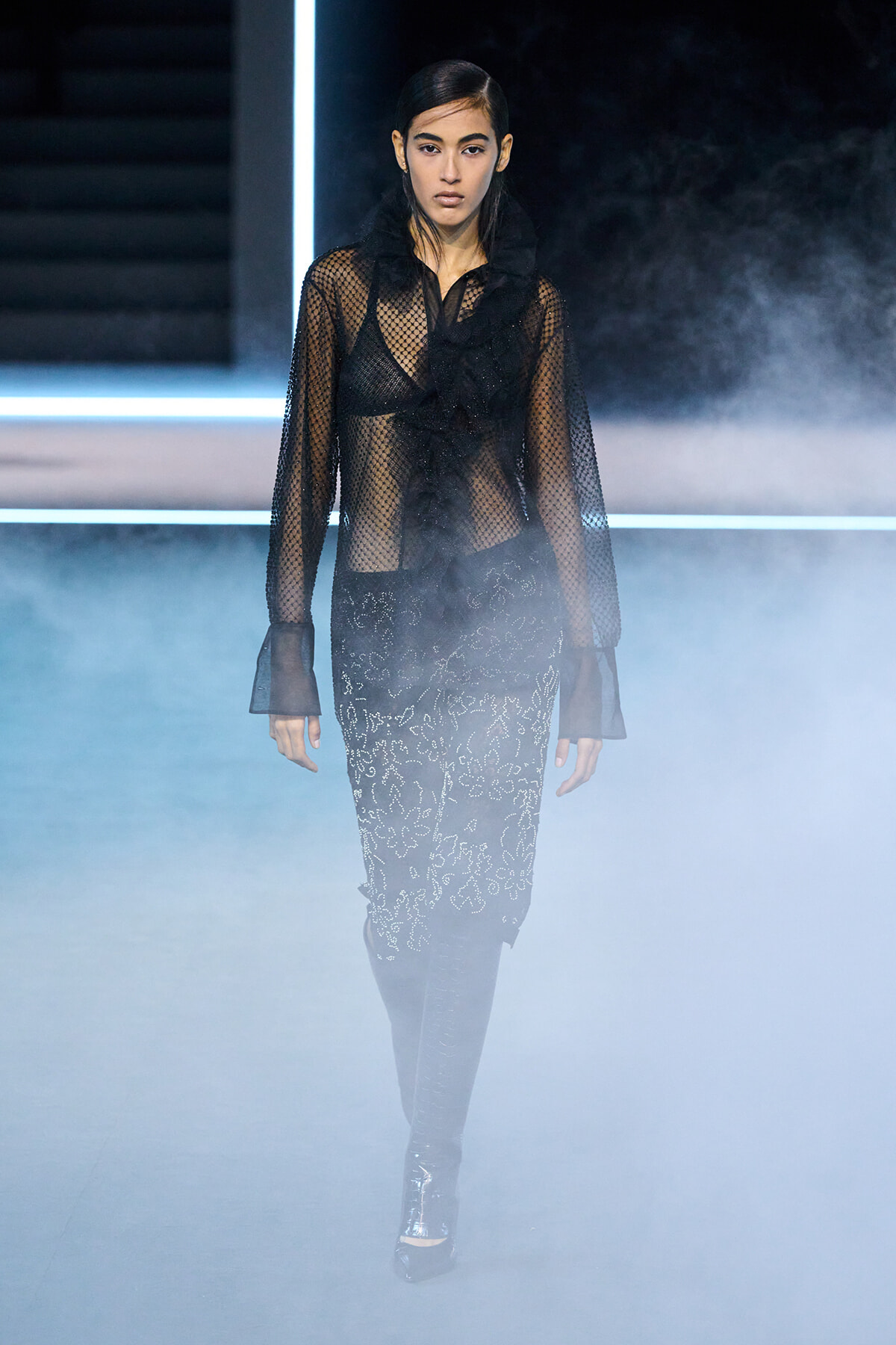 Model walking a fashion runway in a sheer black lace blouse and embroidered skirt, with blue fog and neon light bars in the background.