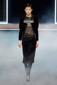 Female model on a fashion runway wearing a black velvet blazer with lace-trim top and black pants, metallic heeled boots, and a chunky choker amid fog.