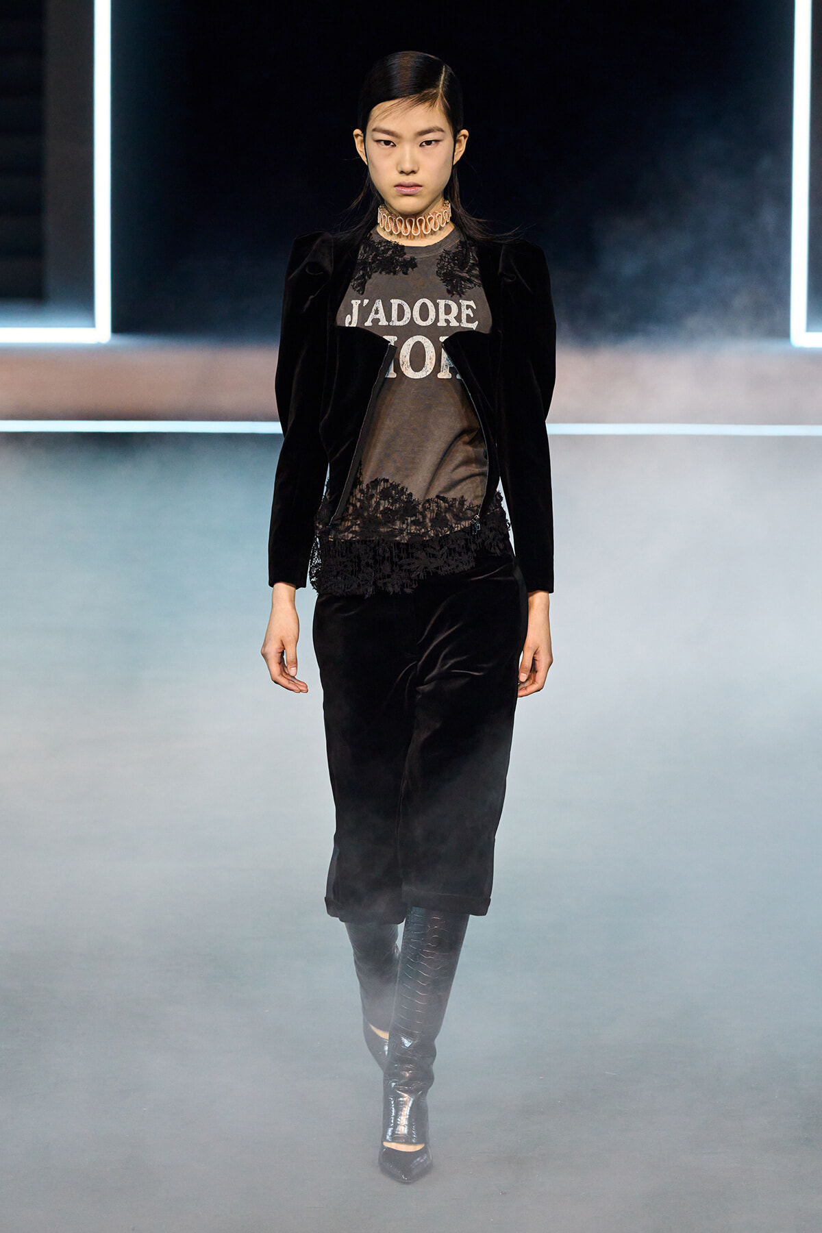Female model on a fashion runway wearing a black velvet blazer with lace-trim top and black pants, metallic heeled boots, and a chunky choker amid fog.