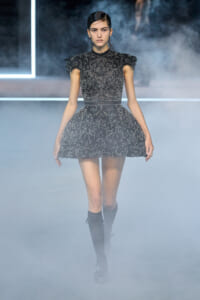 Model walks a smoky runway in a short black lace dress with intricate floral embroidery and puff sleeves, paired with knee-high boots