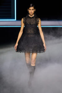 Model walking a runway in a black lattice dress with a fitted bodice and flared skirt, paired with knee-high boots and a choker, amid blue-lit stage and fog.