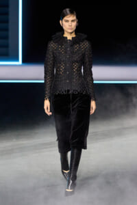 Model walking a runway in a black lace jacket with a high ruffled collar, sheer lattice pattern, paired with velvet wide-leg pants and knee-high boots.